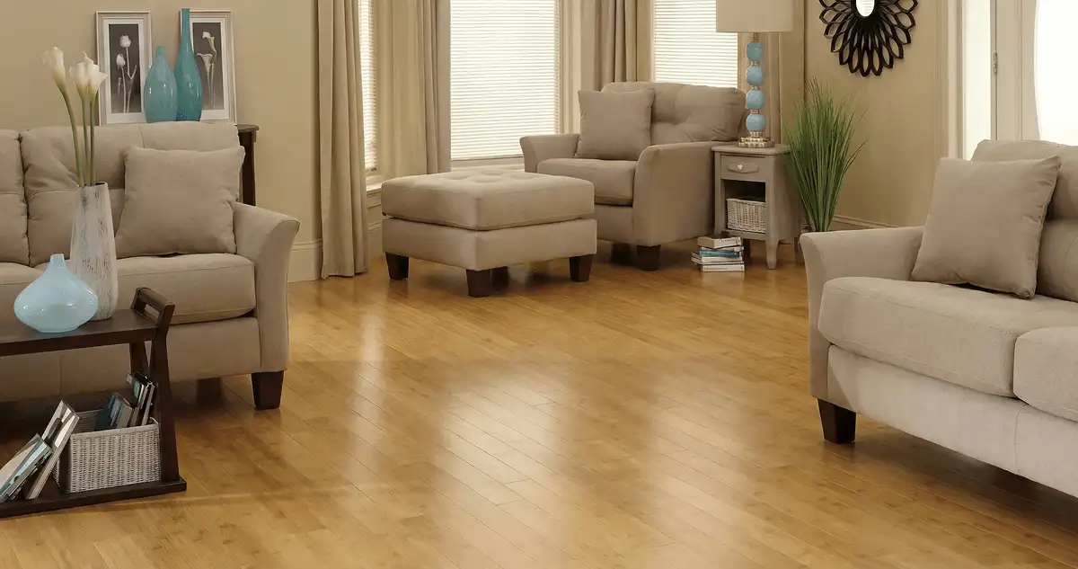 A living room with professionally installed bamboo flooring, beige sofas, a matching ottoman, a side table with a blue lamp, vases, framed pictures, and large windows with blinds and curtains in Fresno, CA